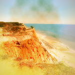 Photographie d'une falaise aux teintes ocres au bord d'une plage de sable fin doré. L'océan bleu azur s'étend à perte de vue.
