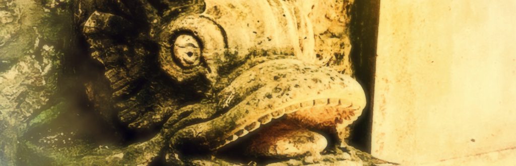 Photographie d'une statue en pierre représentant un triton à la gueule ouverte. La pierre ancienne aux reflets jaunes montre les marques du temps, la mousse le recouvre.