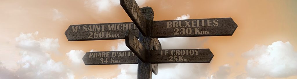 Photographie prise de la falaise au Tréport d'un panneau en bois indiquant les directions aux nuages dans un ciel orangé.
