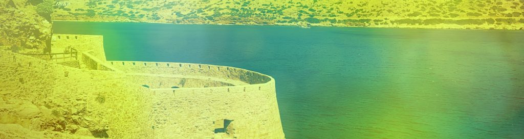 Photographie d'une tour de la forteresse de l'ile de Spinalonga en Crète bordée par les montagne et la mer turquoise.
