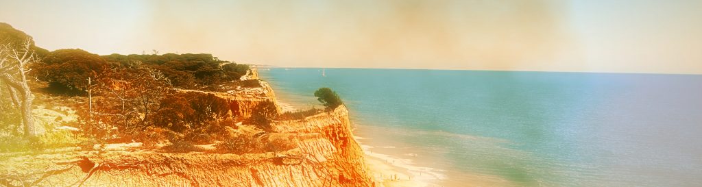 Photographie d'une falaise aux teintes ocres au bord d'une plage de sable fin doré. L'océan bleu azur s'étend à perte de vue.