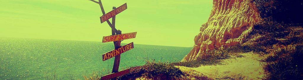 Photographie prise au bord d'une falaise, l'océan bleu en arrière plan. Une pancarte de bois indique des destinations.