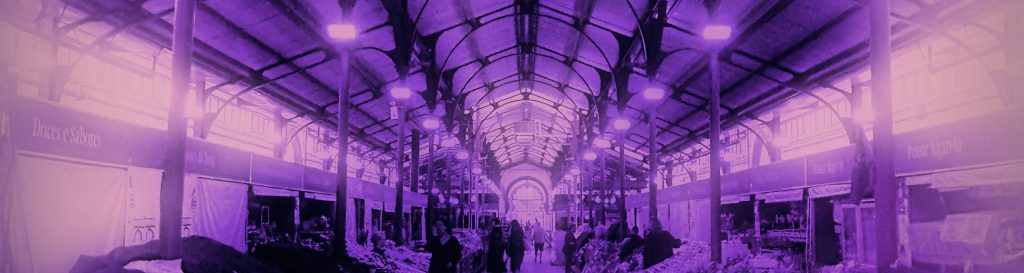 Photographie d'une allée du marché de Loulé. Le toit est soutenu par une ossature métallique. Au bout de l'allée, la porte illuminée par le soleil forme une arche.
