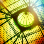 Photographie représentant le toit d'un bâtiment, une verrière faite de vitre et de poutres en fer. La lumière se reflète sur les vitres provocant un arc en ciel de lumière.